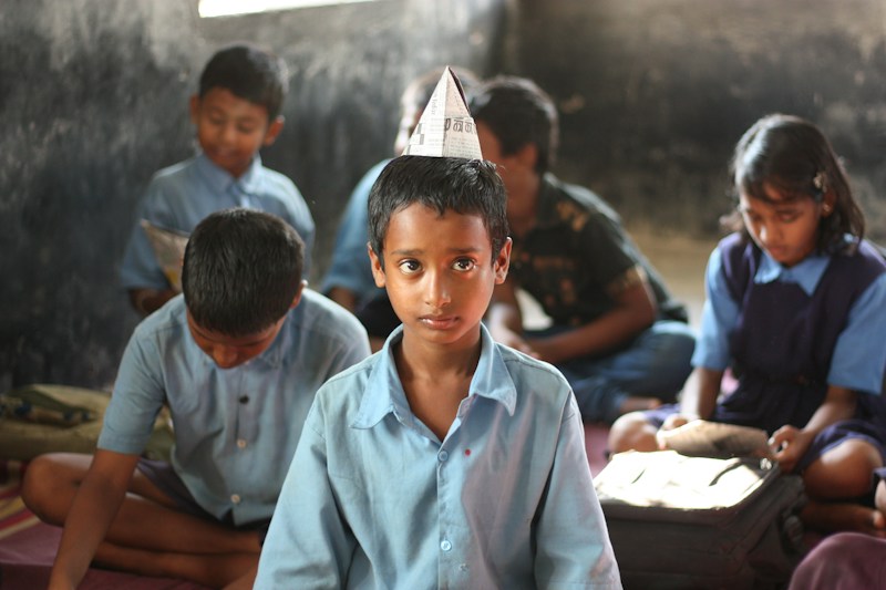 A boy in a paper hat sits in the front row of his classroom