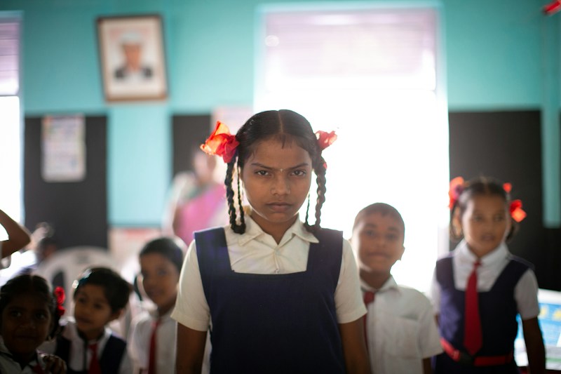 A schoolgirl in uniform with plaited hair stands confidently in her classroom