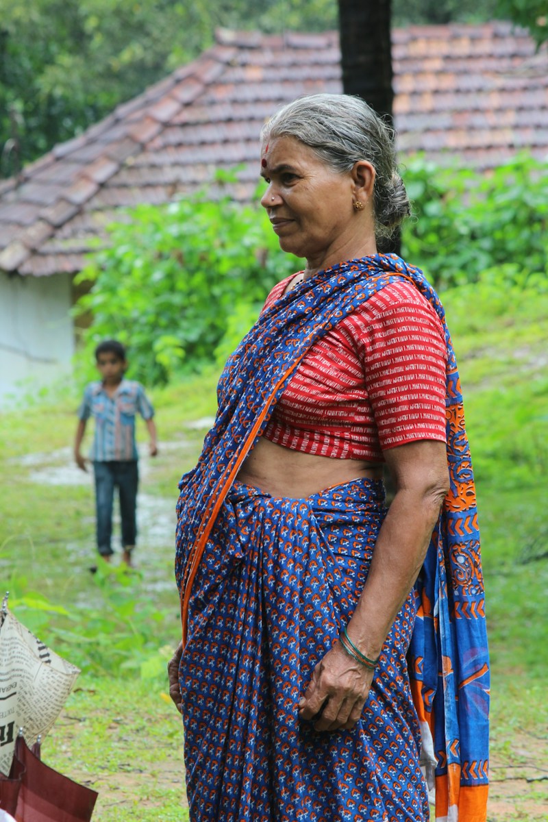 An older woman in a patterned sari stands in front of her village home, a child playing behind her